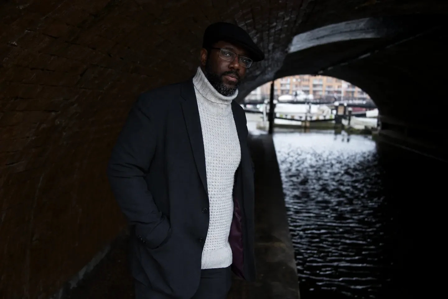 Dr Gyles Morrison by a canal under a brick arch, wearing a dark cap and light cable-knit jumper